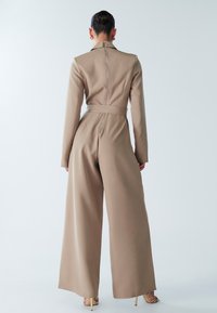 Bēša kombinezons ar piegulošu augšdaļu, garām piedurknēm un platām biksēm. Aprīkots ar augstu apkakli un aizmugures rāvējslēdzēju, piejaucēts ar atbilstošu jostu.