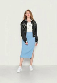 Casaco bombardeiro preto com riscas brancas e lettering "Gensdale", t-shirt branca, saia midi azul canelada, e sapatilhas brancas.