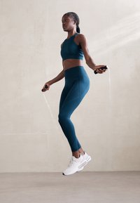 Teal sporttop en high-waisted legging van een gladde stof, gecombineerd met witte sportschoenen, terwijl er binnen met een springtouw wordt geoefend.