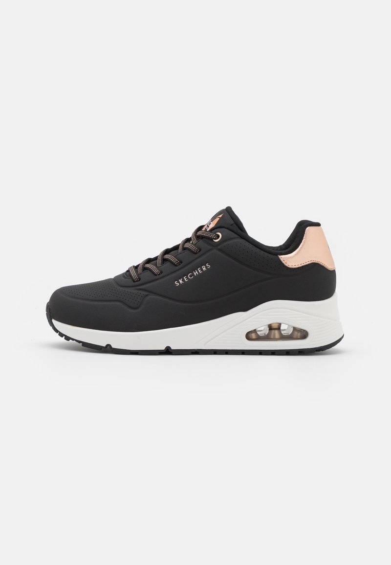 Baskets basses Skechers noires avec semelle blanche, accent talon en or rose, lacets noirs et blancs, et coussin d'air visible sur le panneau latéral de la semelle.