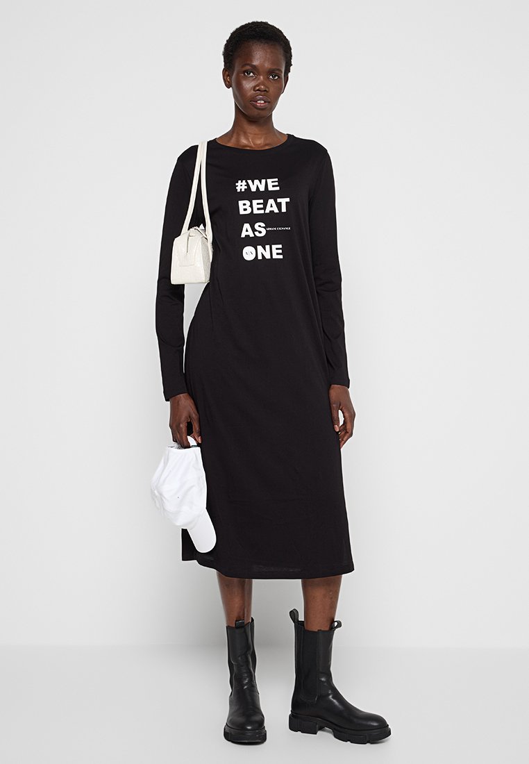 Abito lungo nero a maniche lunghe con testo bianco "#WE BEAT AS ONE," caratterizzato da una vestibilità comoda. Abbinato a stivali neri e una piccola borsa bianca.