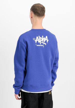 Man wearing blue sweatshirt with white "Alpha Industries" logo on back, paired with black pants against plain white background.