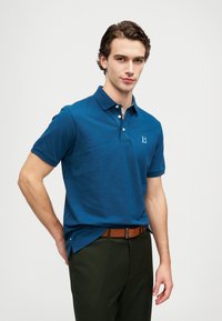 Blue polo shirt made of breathable fabric, with a collar, three buttons, and a small logo on the chest, paired with green pants and a brown belt.
