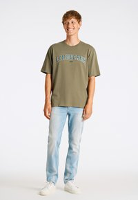 Olivgrön T-shirt med "LINDBERGH 1927" tryckt i blått, parat med ljusblå jeans och vita sneakers. Avslappnad passform och kort ärm.