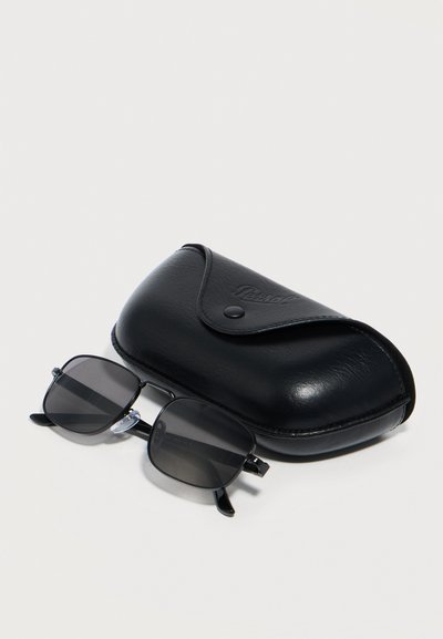Μαύρα ορθογώνια γυαλιά ηλίου με σκουρόχρωμους φακούς τοποθετημένα δίπλα σε μια κλειστή μαύρη δερμάτινη θήκη πάνω σε λευκή επιφάνεια.