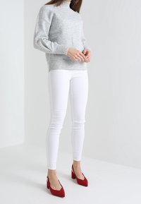 Suéter gris de punto con un cuello alto, jeans skinny blancos y zapatos de tacón rojos, con tacones de bloque y un detalle de cremallera.