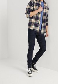 Rutig flanellskjorta i marinblått, grönt och orange över en blå t-shirt, ihop med mörka tajta jeans och svarta högskurna sneakers.