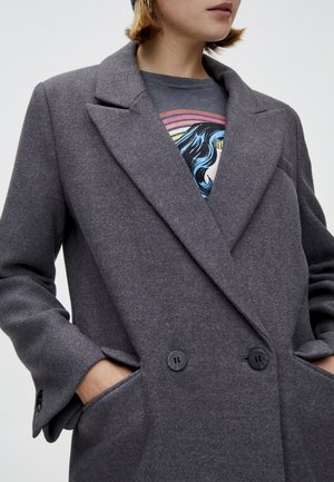 Personne portant un manteau gris à double boutonnage, les mains dans les poches, sous lequel un t-shirt graphique avec des cheveux bleus et des rayures arc-en-ciel est partiellement visible.