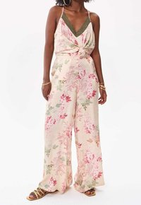 Blommig jumpsuit i ljusrosa tyg med grönt spetsband. Har vida ben och en knuten detalj fram. Mjuk, lätt textur.