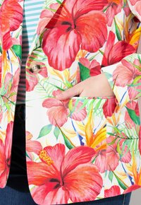 Blumenblazer mit weißem Hintergrund, der lebhafte rote Hibiskusblumen und grüne Blätter zeigt. Enthält eine Tasche und ein Knopfdetail.
