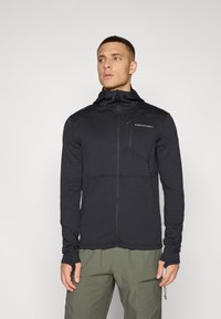 Giacca nera con zip e finitura texture, dotata di cappuccio, maniche aderenti e tasca sul petto. Abbinata a pantaloni verde oliva.