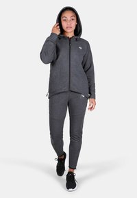 Sivá mikina na zips s kapucňou a šnúrkami, skombinovaná s ladiacimi tapered teplákmi. Obe sú vyrobené z mäkkej látky. Čierne tenisky dokončujú vzhľad.