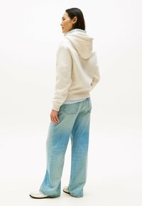 Beige Kapuzenpullover mit weicher Textur, kombiniert mit hellblauen weit geschnittenen Jeans. Modell wird von hinten vor einem schlichten Hintergrund gezeigt.