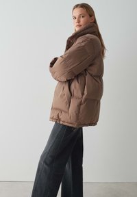 Giacca a vento marrone con colletto alto e foderato in pelliccia. Presenta una vestibilità ampia, texture trapuntata e orlo elasticizzato. Abbinata a jeans larghi neri.