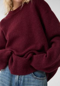 Pull en maille bordeaux à coupe décontractée, doté d'une texture douce et de poignets retroussés, associé à un jean en denim bleu.