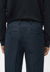 Rückansicht einer Person, die dunkelblaue, maßgeschneiderte Hosen mit geknöpften Gesäßtaschen und ein schwarzes, langärmeliges Oberteil trägt, das in die Hose gesteckt ist.