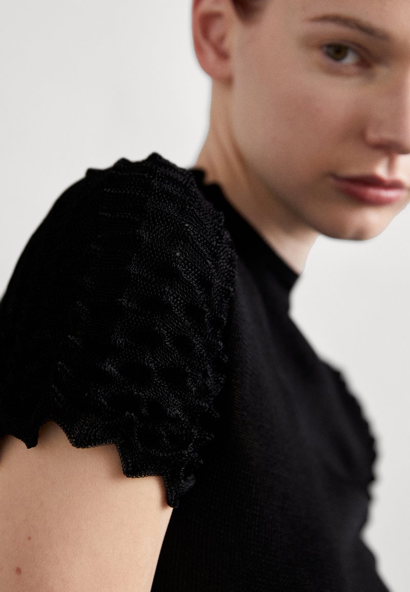 Top de punto negro con mangas texturizadas y con volantes. Primer plano que muestra el detalle del patrón de la tela y el escote. Material del cuerpo suave, diseño ajustado.