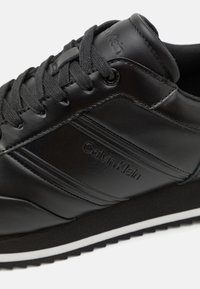Zapatilla deportiva de cuero negro con acabado texturizado, que presenta un destacado logo de Calvin Klein, diseño con cordones y una suela intermedia blanca en contraste.