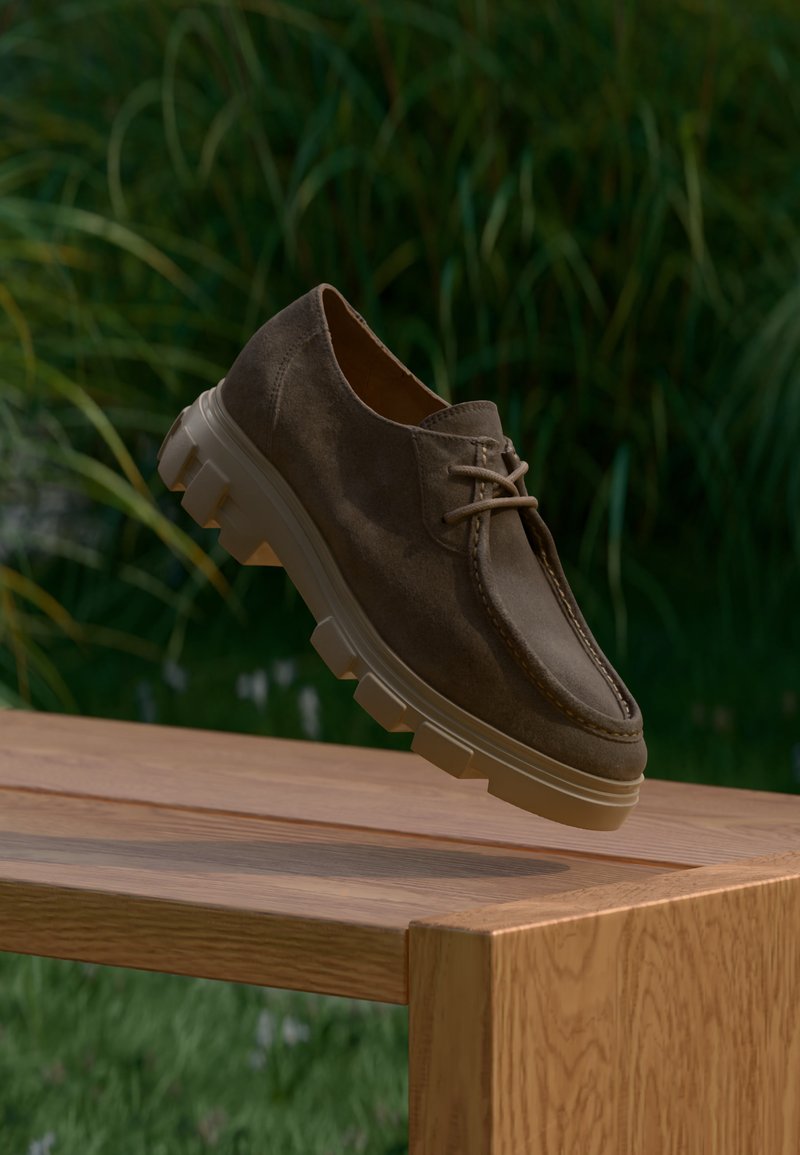 Chaussure à lacets en daim marron avec semelle beige épaisse et nervurée, flottant au-dessus d'un banc en bois en plein air avec un fond de feuillage vert.