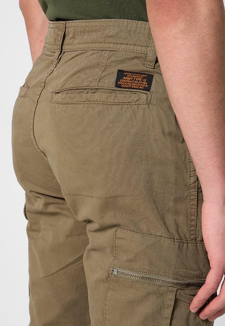 Primer plano de una persona que lleva pantalones cargo verde oliva con etiqueta en el bolsillo trasero y bolsillo lateral con cremallera, mano apoyada cerca del muslo.