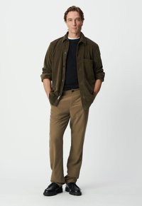 Olivenfarbendes Cordhemd, schwarzes T-Shirt, beige Hose und schwarze Schuhe. Das Hemd hat eine Brusttasche; die Hose ist tailliert und hat einen Knopfverschluss.