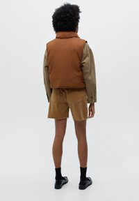 Brown padded vest over a green shirt, paired with tan shorts. Notable features include a high collar and stitching details.