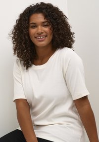 Light cream short-sleeve top in ribbed texture with a round neckline, featuring rolled sleeves and a relaxed fit. Model is seated.