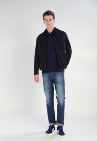 Črna suknjič iz nubuk usnja s pritisnimi gumbi, mornarsko modra polo majica, svetlo modri kavbojke in mornarsko modri športni copati z belimi detajli. Model stoji obrnjeno naprej.