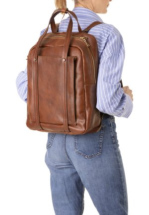 Femme portant un sac à dos en cuir marron avec deux poignées, vêtue d'une chemise rayée bleue et d'un jean bleu, vue de dos.