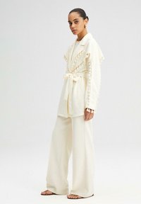 Chaqueta color crema con bordes con flecos, atada en la cintura, combinada con pantalones de pierna ancha también color crema. Ambas prendas están hechas de una tela suave.