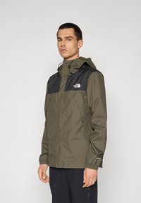 Olijfgroene waterdichte jas met zwarte accenten op de schouders, verstelbare capuchon en elastische manchetten. Beschikt over het North Face-logo op de borst.