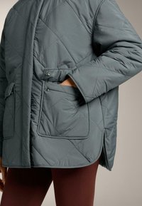 Krojena siva jakna s džepovima na kopčanje koju nosi osoba s jednom rukom u džepu i vidljivim tamnocrvenim hlačama.