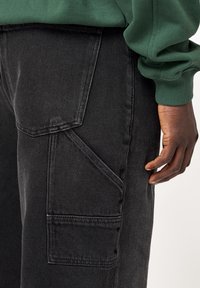 Schwarze Denim-Cargohosen mit mehreren Taschen. Der Stoff hat eine glatte Textur und ein verblasstes Finish. Ein grüner Pullover ist teilweise sichtbar.