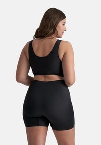 Donna con capelli castani della lunghezza delle spalle, vestita con un top corto senza maniche nero e pantaloni corti neri a vita alta, vista di spalle.