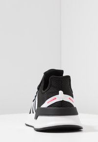 Svart och vit atletikssko med mesh-överdel, vit sula och röd sömdetalj vid hälen, visad från en bakvinkel på en vit yta.