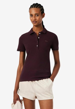 Mujer con el cabello trenzado, vistiendo una camiseta tipo polo morado oscuro y pantalones cortos beige claro, de pie con una mano en el bolsillo y la otra sosteniendo una tela.