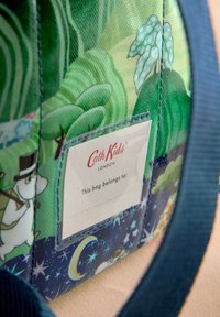 Zelena platnena torba z domiselnimi motivi dreves in likov, z velikim pravokotnim etiketom, ki navaja lastništvo. Modra ročaja.