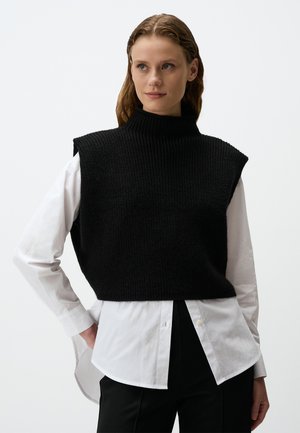 Zwarte gebreide mouwloze trui met een hoge hals, gedragen over een wit shirt, met een ribbelstructuur en een cropped design.