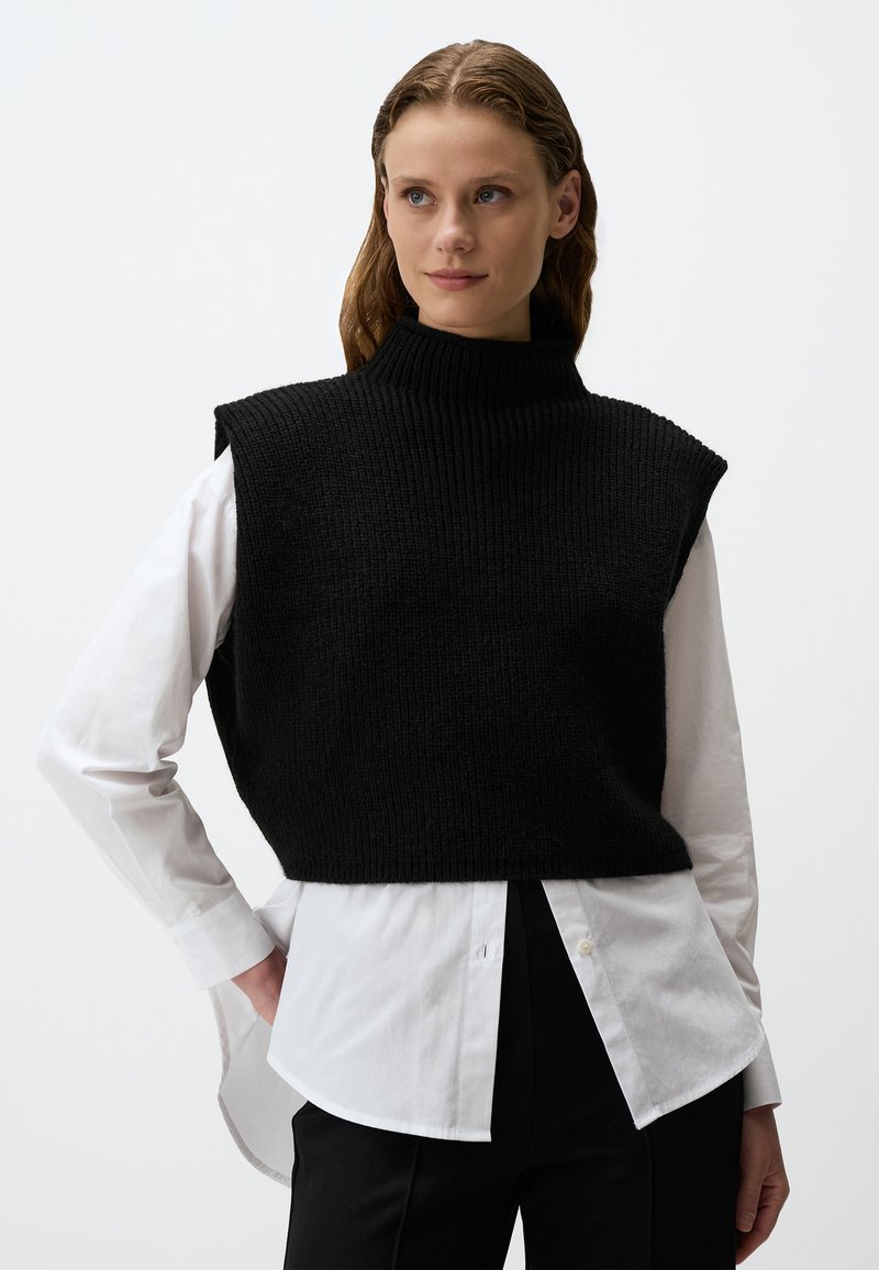 Zwarte gebreide mouwloze trui met een hoge hals, gedragen over een wit shirt, met een ribbelstructuur en een cropped design.