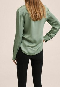 Camicia lunga in satin verde con orlo arrotondato, polsini aderenti e una trama liscia. Abbinata a pantaloni neri. Vista posteriore.