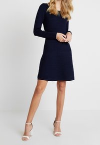 Robe à manches longues bleu marine en tissu texturé, avec un corsage ajusté et une jupe évasée. Associée à des talons blancs à lanières pour contraster.