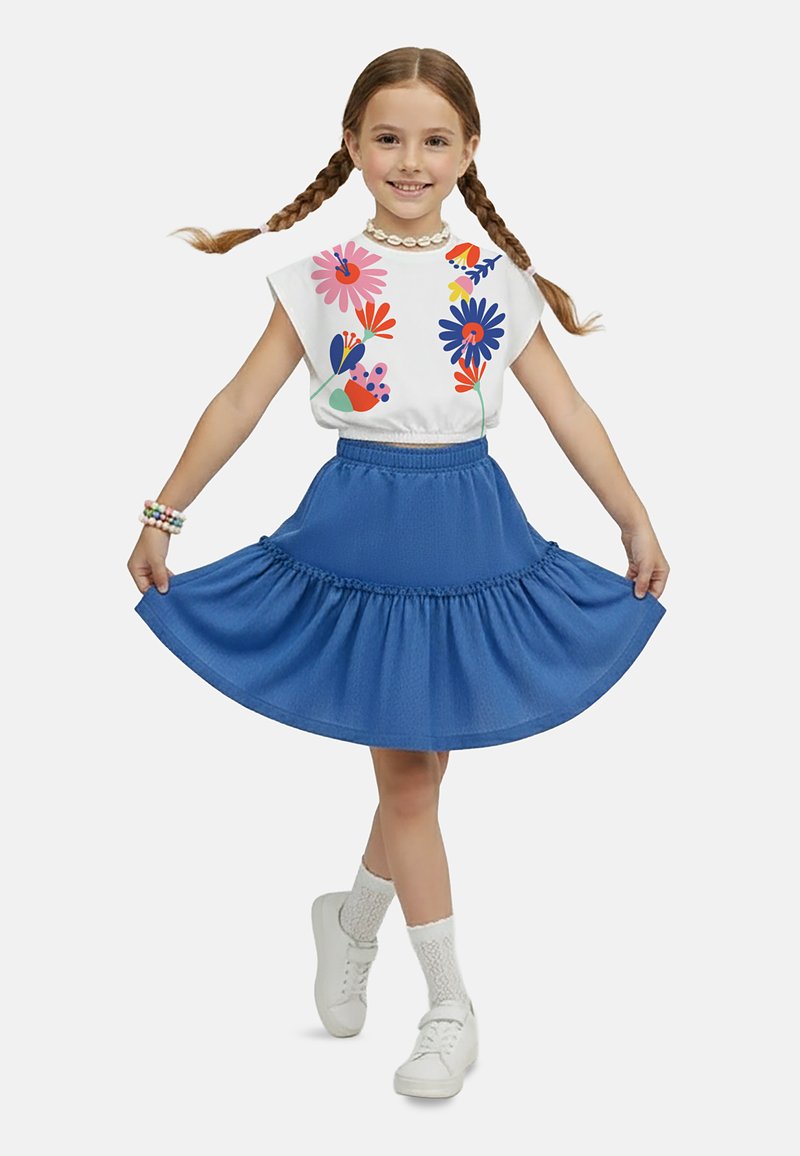 Ung pige med fletninger, iført en hvid blomstret top, blåt skørt, hvide sokker, sneakers og halskæde med muslinger, smilende og holder sin nederdel.