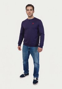 Jonge man staat in een marineblauw shirt met lange mouwen, een blauwe spijkerbroek en marineblauwe sneakers met witte veters voor een plain witte achtergrond.