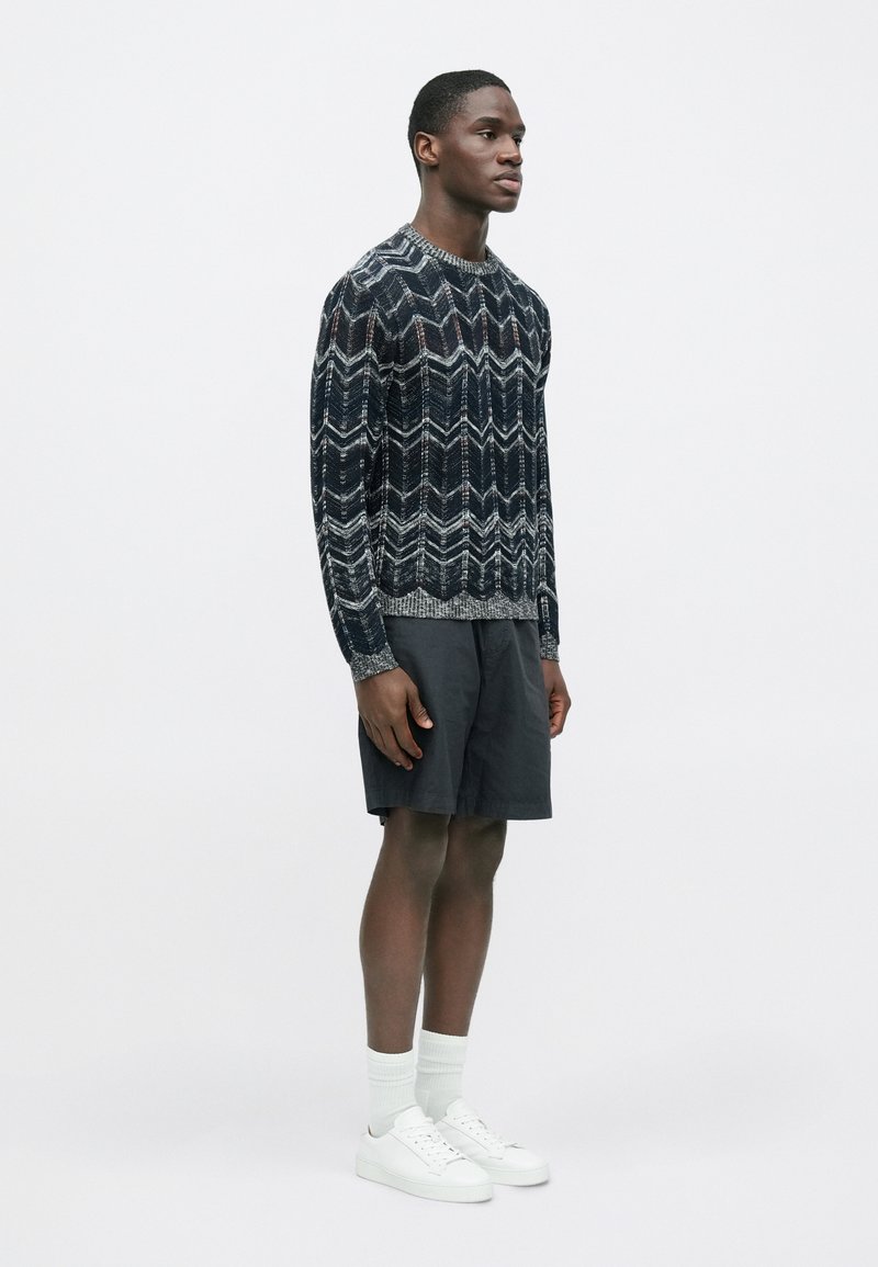 Pull en maille à motif sombre avec un design en chevrons, associé à un short foncé et des baskets blanches portées avec des chaussettes côtelées à hauteur de mollet.