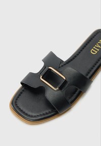 Black flat sandal featuring a wide strap with gold-tone buckle detail, textured upper, and a flat sole with a slight curve.