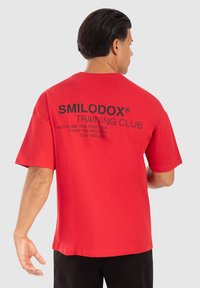 Rød bomulds t-shirt med afslappet pasform, korte ærmer og sort trykt tekst på ryggen, herunder "SMILODOX® TRAINING CLUB" og motiverende sætninger.