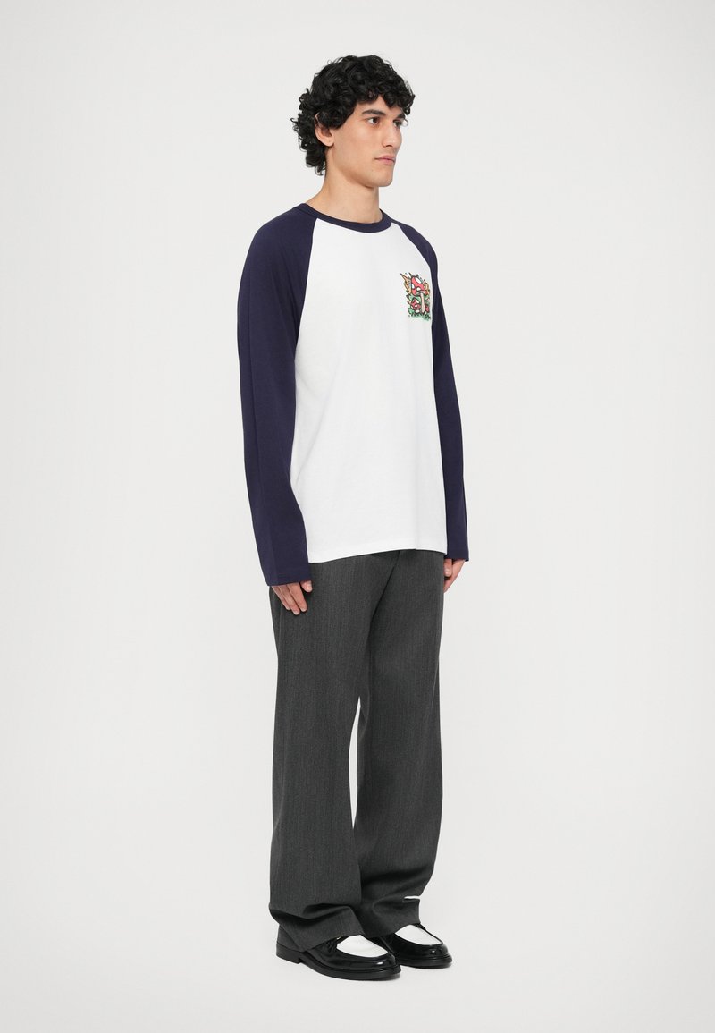 T-shirt blanc avec des manches raglan bleu marine, arborant un graphique coloré sur la poitrine gauche. Assorti à un pantalon ample gris foncé et des chaussures noires.