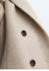 Beige frakkestof med en blød tekstur, der har mørke knapper og en reversdesign. Nærbillede af frakkens detaljer og syninger.