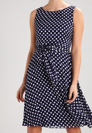 Robe bleu marine sans manches à pois blancs, taille cintrée nouée en un nœud, portée par une personne debout devant un fond uni.