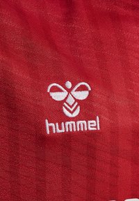 Tessuto rosso con un logo ricamato bianco che presenta forme simmetriche e la scritta "hummel", caratterizzato da una texture liscia.
